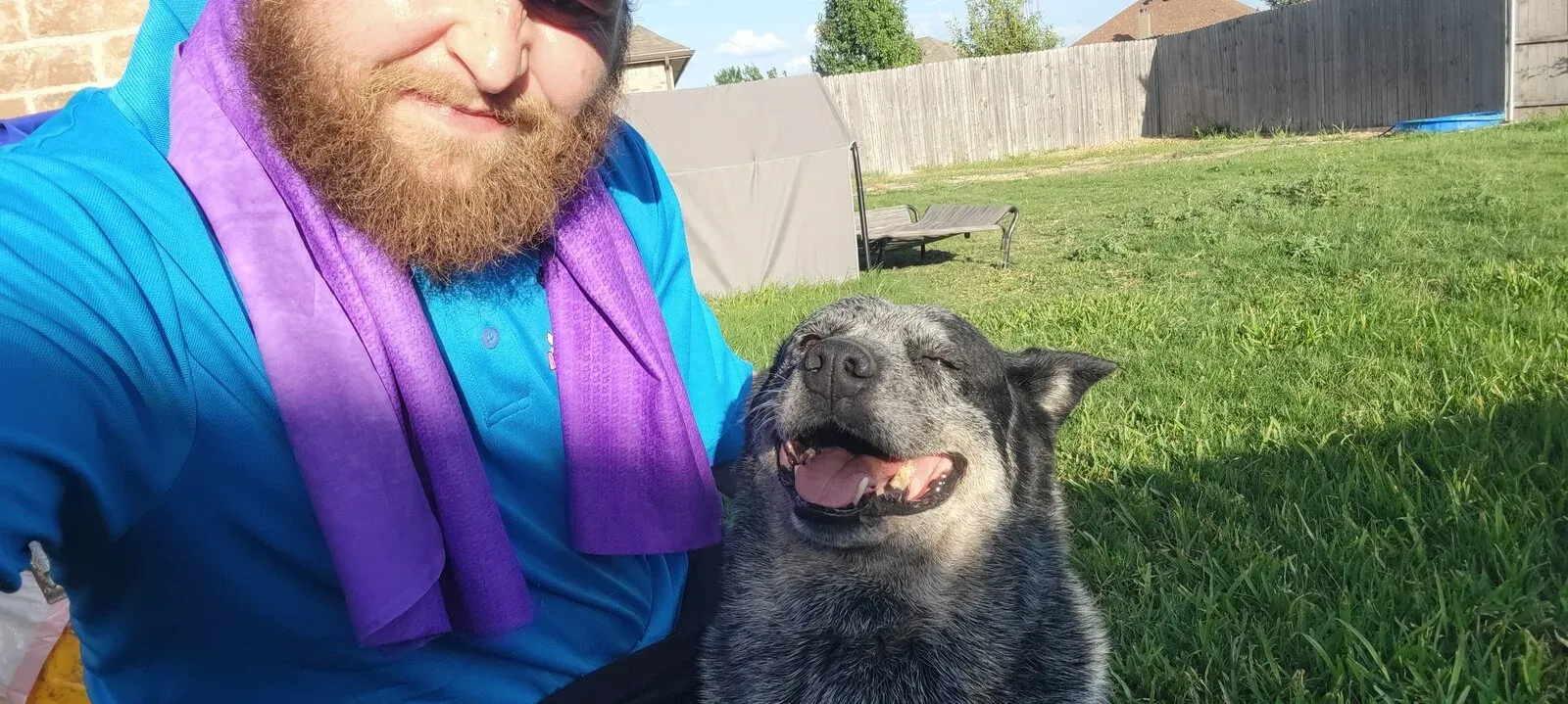 A happy blue heeler in a backyard next to Brandon in his blue Booty Scoopy polo