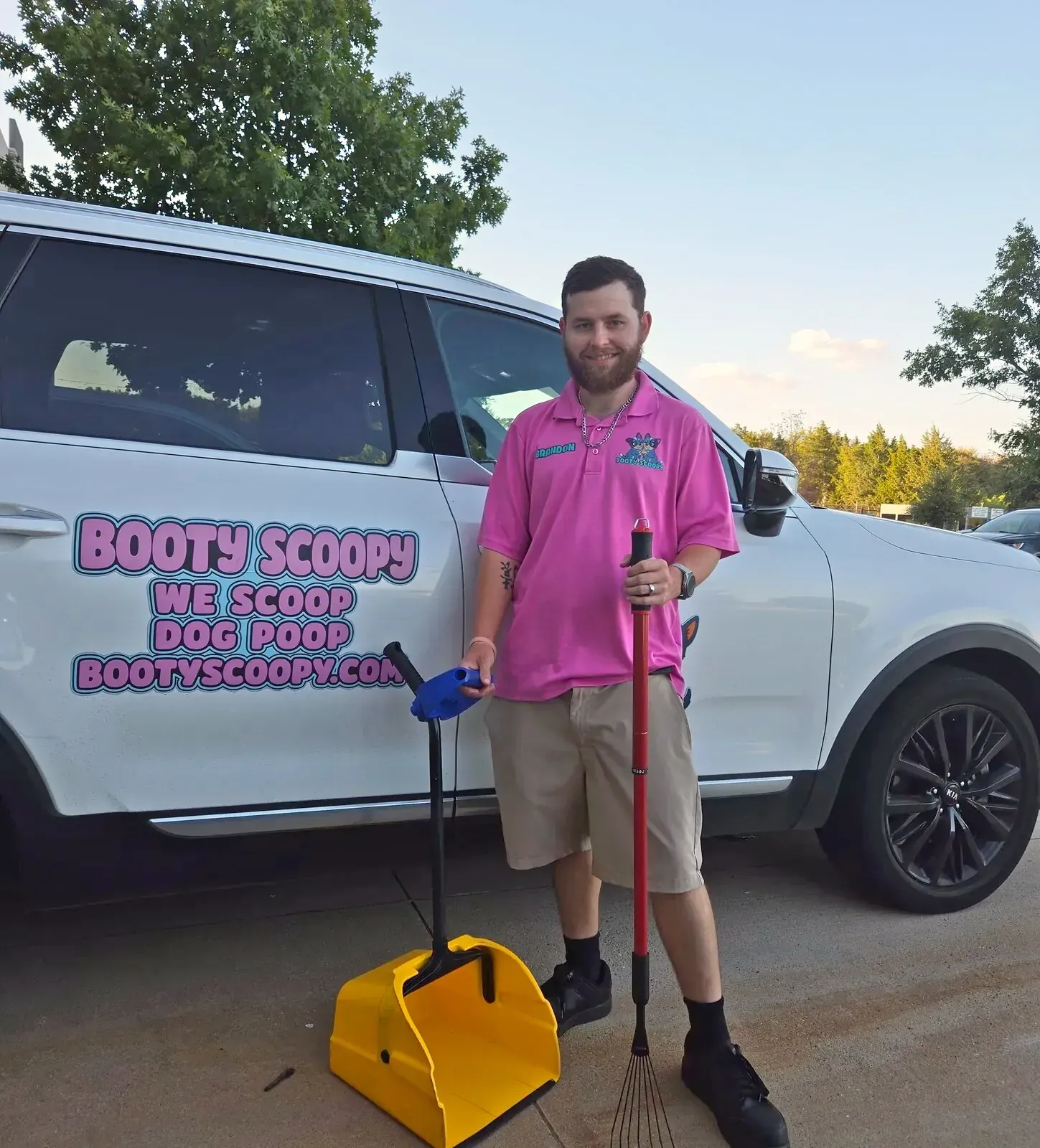 Brandon in his pink Booty Scoopy polo with his rake and yellow scoop pan beside the lettered Booty Scoopy SUV